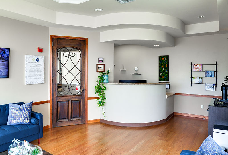 Reception area with a wooden door with wrought iron details, curved white reception desk, blue chairs with patterned pillows, and wall decorations including plants and framed art.