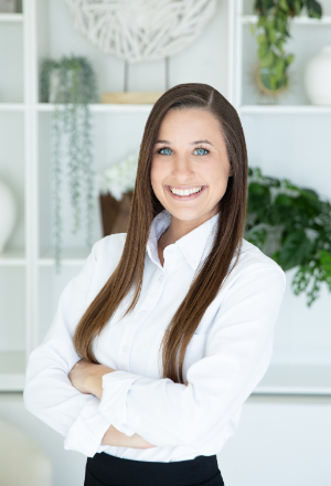 Smiling woman with long brown hair, wearing a white shirt, standing with arms crossed in front of a decorated white shelf.