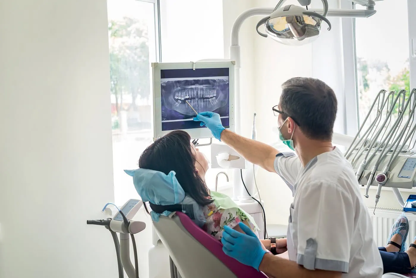 Dentist wearing gloves and mask showing a dental X-ray to a patient seated in a dental chair.