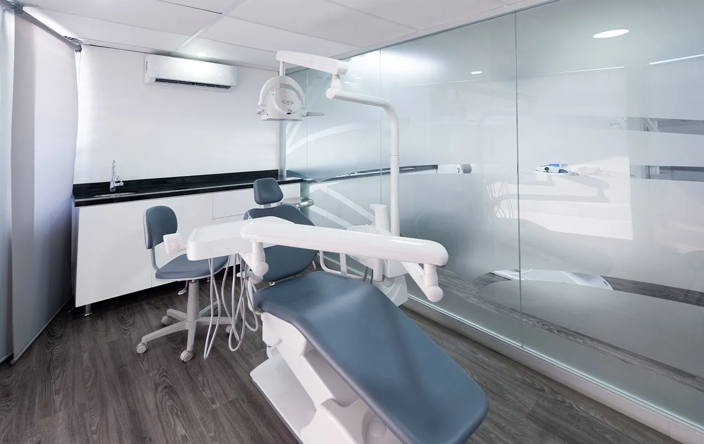 Modern dental clinic room with a dental chair, dentist stool, overhead light, white cabinets, and glass partition.