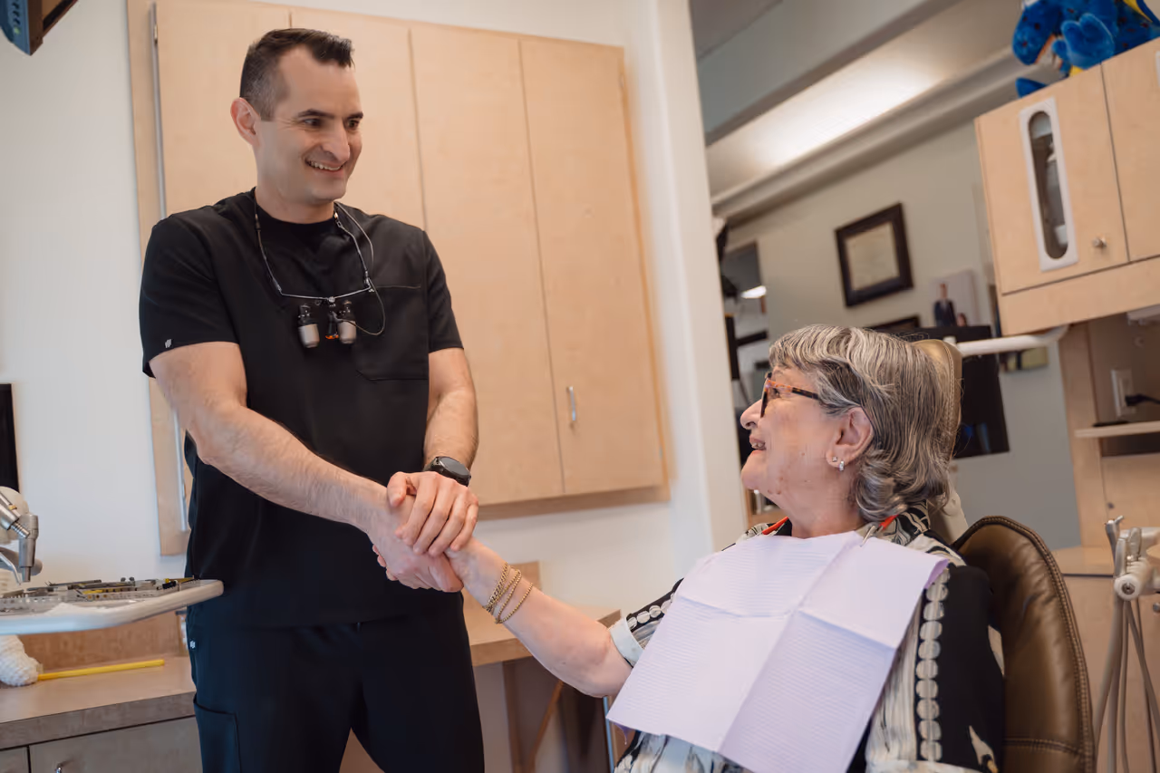 Smiling dentist shaking hands with an elderly female patient sitting in a dental chair.