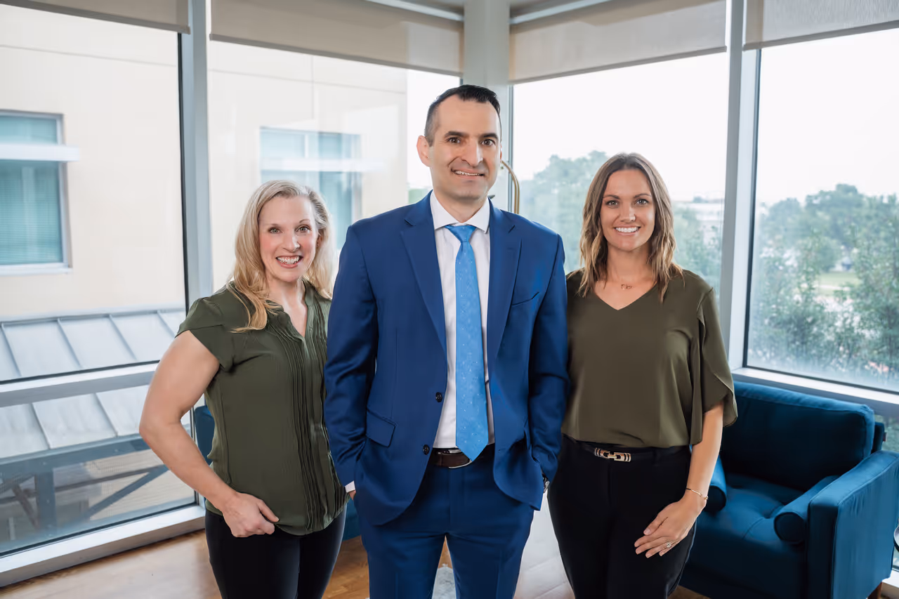 Three smiling professionals standing in an office with large windows, two women wearing olive green tops and a man in a blue suit and tie.