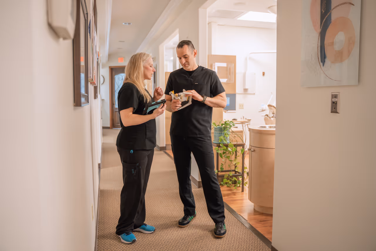 Two dental professionals in black scrubs discussing a dental model in a clinic hallway.
