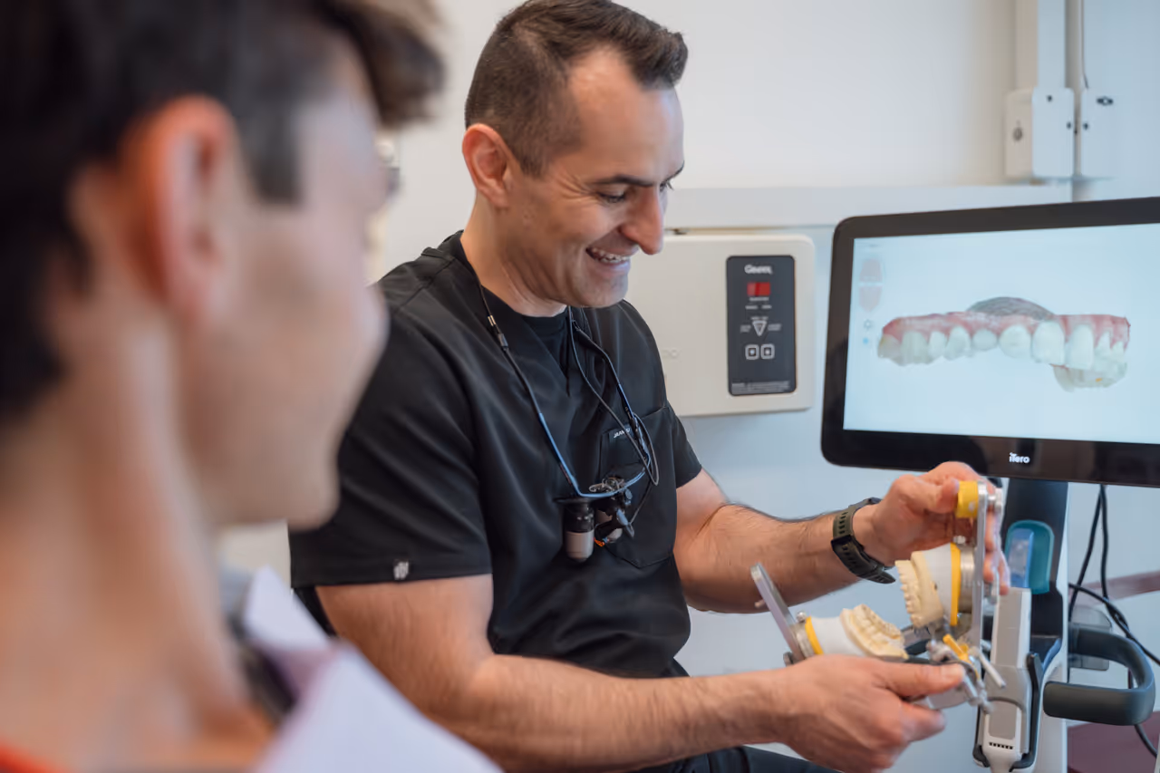 A smiling dentist in black scrubs holding a dental model with a computer screen displaying a 3D image of teeth in the background.