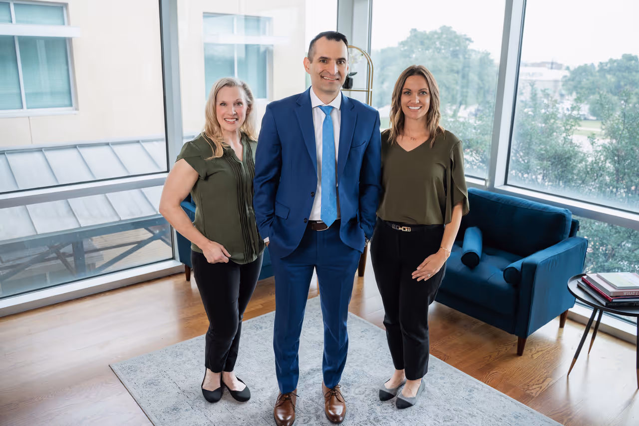 Two women and one man standing and smiling in a bright modern office with large windows and blue armchairs.