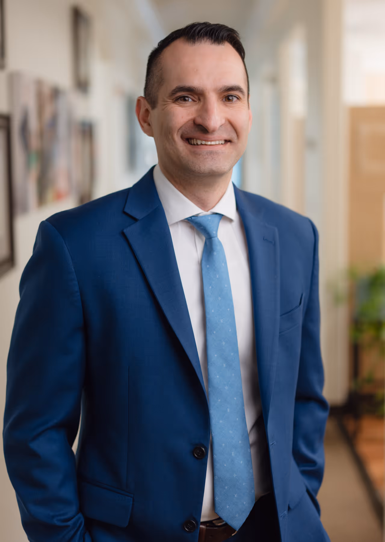 Smiling man in a blue suit, white shirt, and light blue tie standing indoors with blurred background.