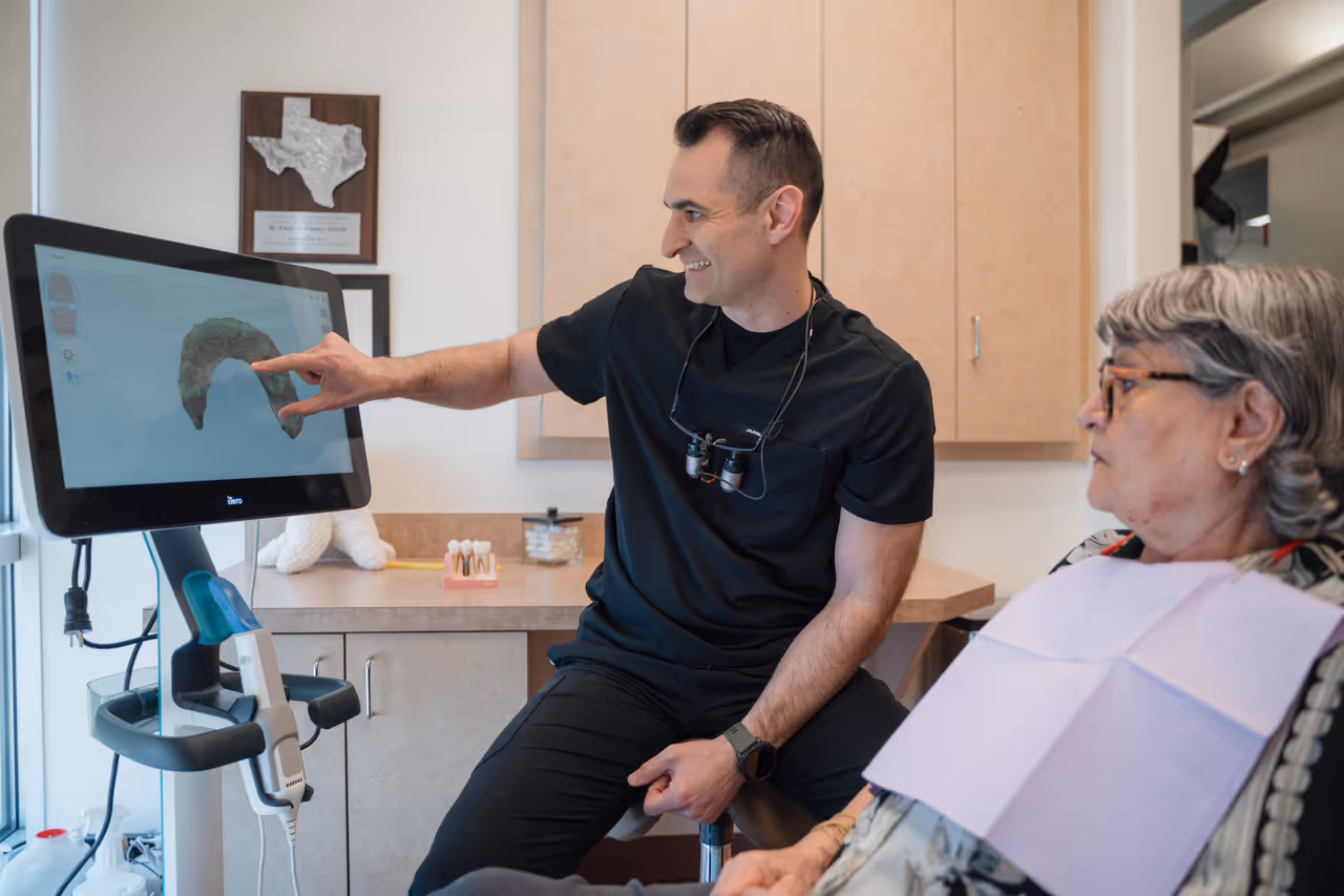 Dentist explaining a digital dental scan on a monitor to a seated elderly female patient in a dental office.