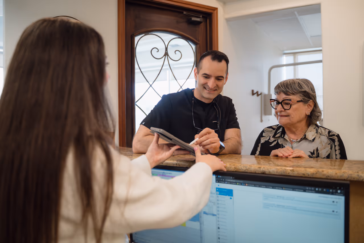 Man and older woman at reception desk interacting with receptionist holding a tablet.