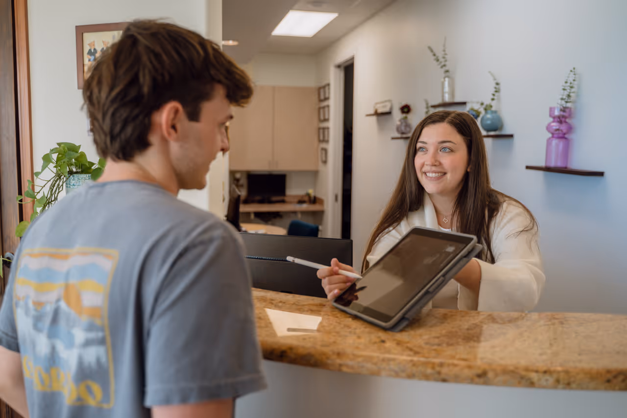 Smiling woman at reception desk showing a tablet screen to a young man wearing a gray t-shirt.