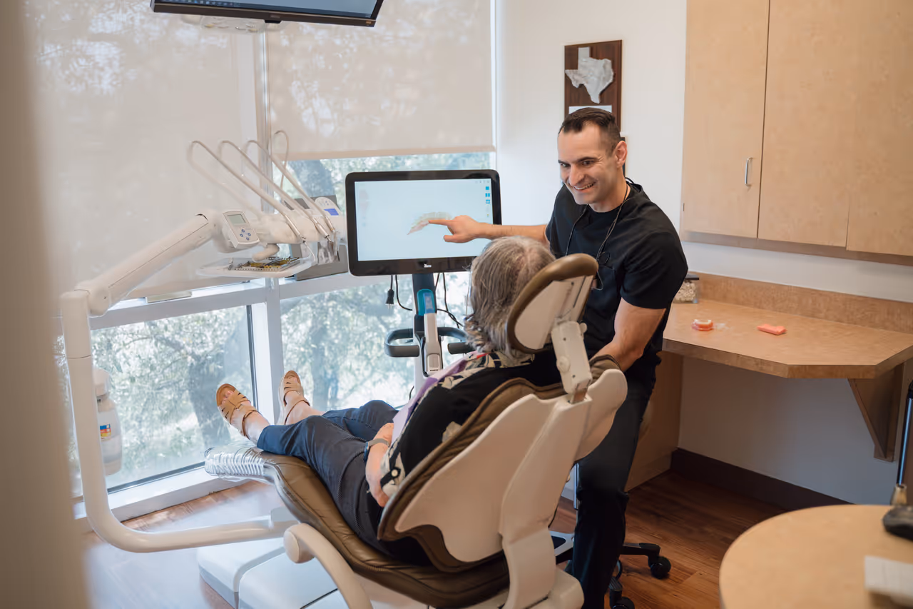 Dentist showing a digital dental scan on a monitor to a patient seated in a dental chair in a bright clinic room.