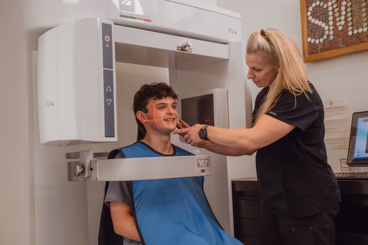 Dental technician adjusting a smiling male patient's head in a dental X-ray machine with red laser alignment lines.