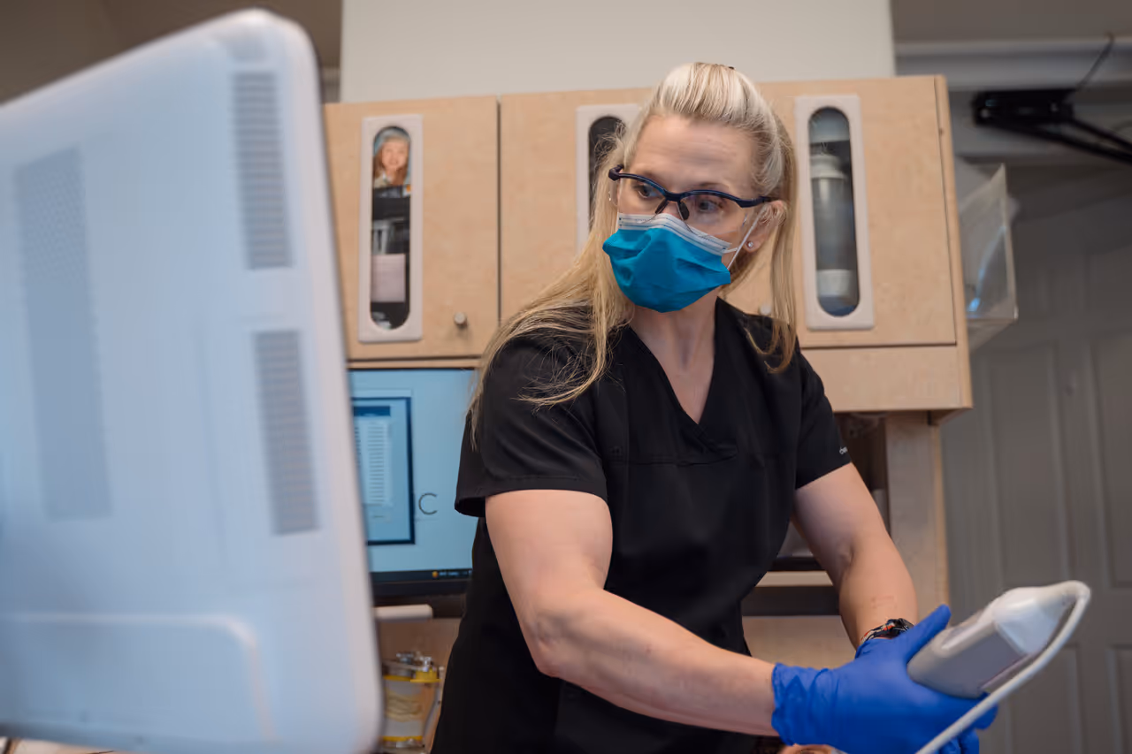 Healthcare professional in black scrubs, wearing protective glasses, blue mask, and blue gloves, operating medical equipment in a clinic.