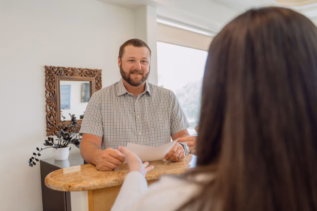 Man in a checkered shirt smiling and receiving a paper from a woman across a marble reception desk.