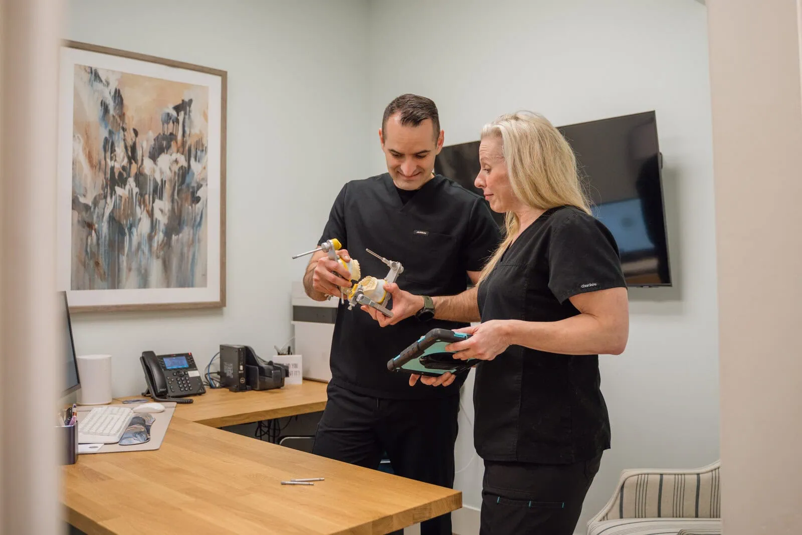 Two dental professionals in black scrubs discuss a dental model and tablet in an office with a wooden desk and wall art.