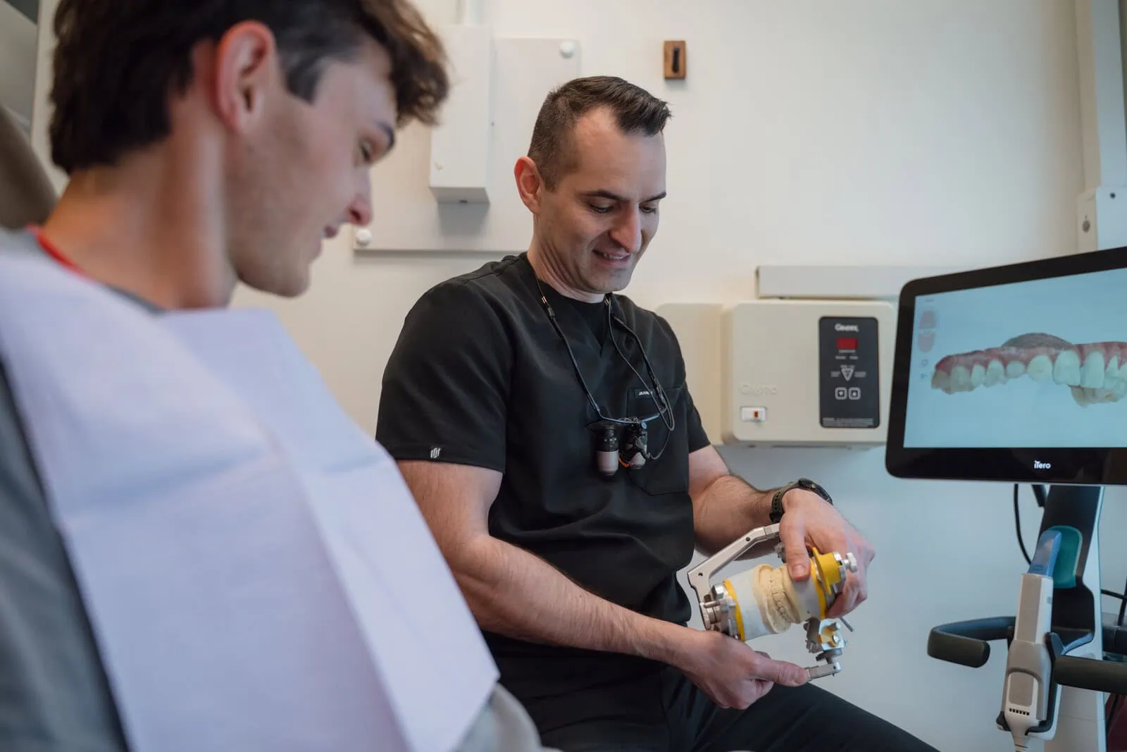 Dentist showing a dental mold to a patient next to a screen displaying a digital dental image.
