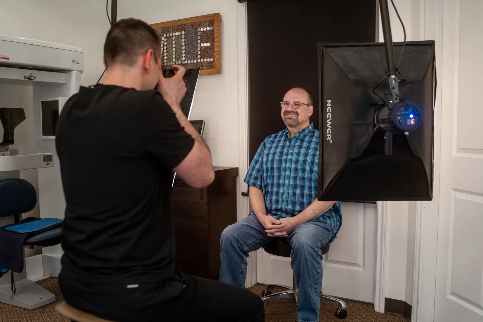 A photographer in a black shirt takes a photo of a smiling man wearing glasses and a blue plaid shirt sitting on a rolling stool in a studio with softbox lighting.